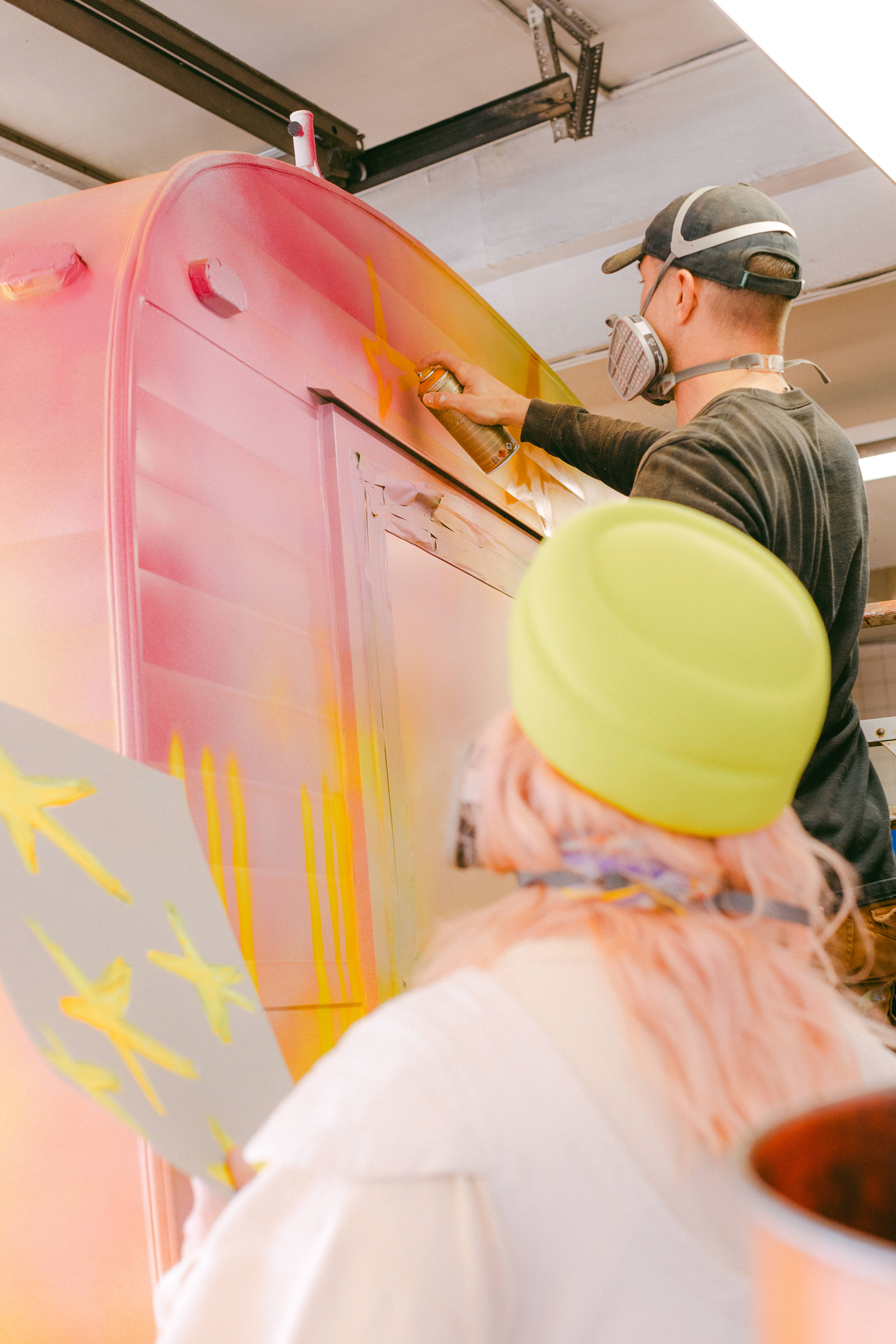 Person spray painting a large pink and yellow mural on a wall.