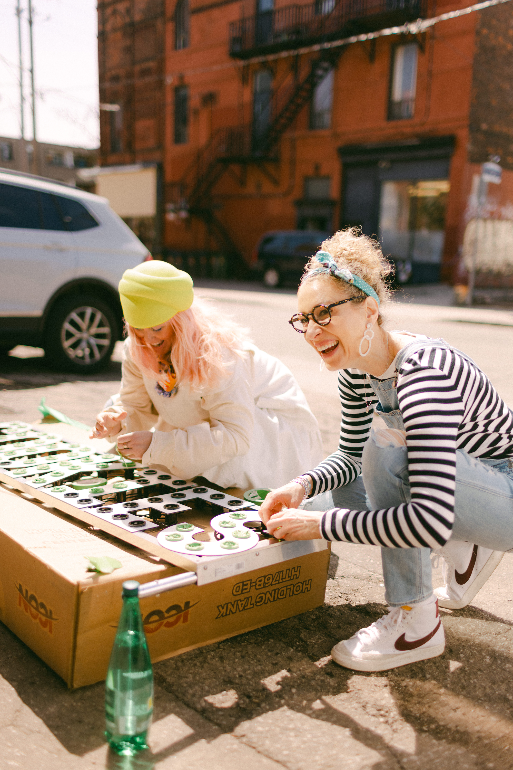 Two women unloading items from a cardboard box on a street with a building in the background.