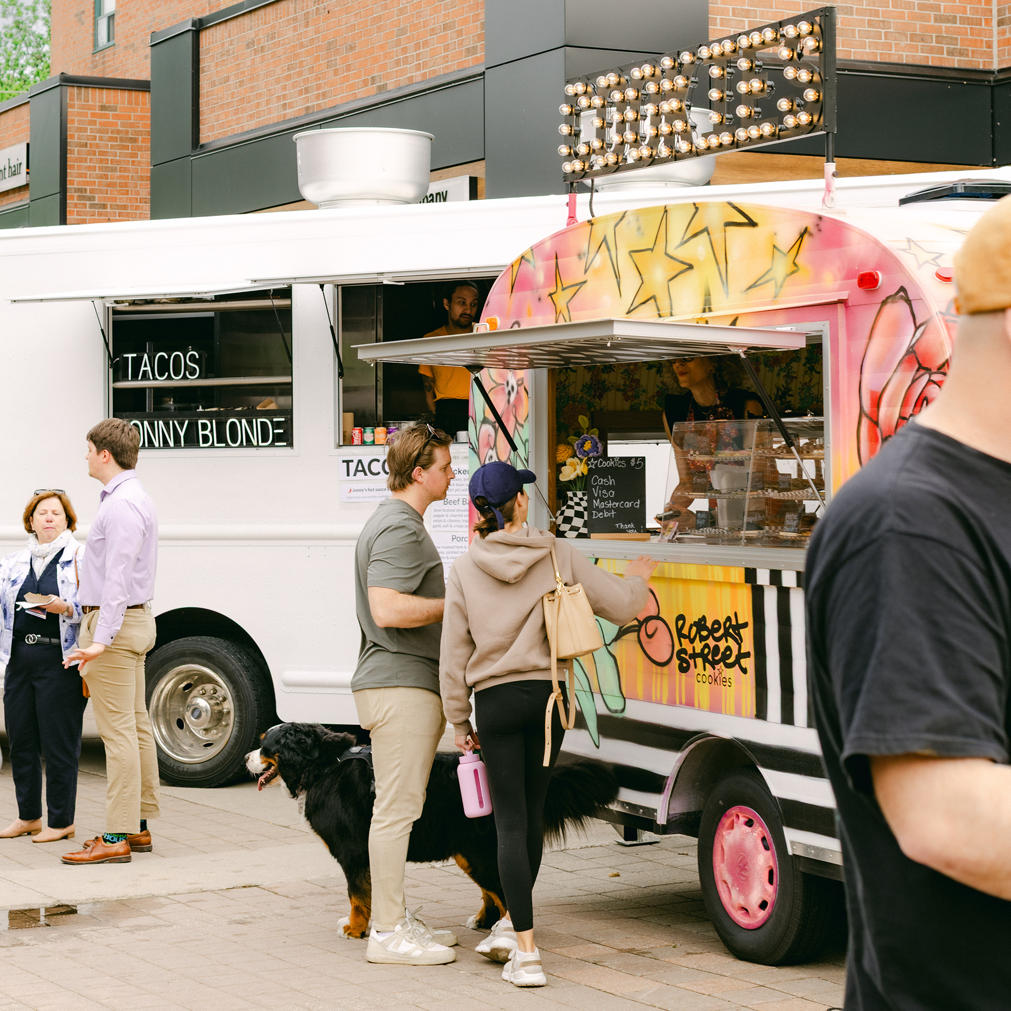 People and a dog standing in front of food trucks on a street.